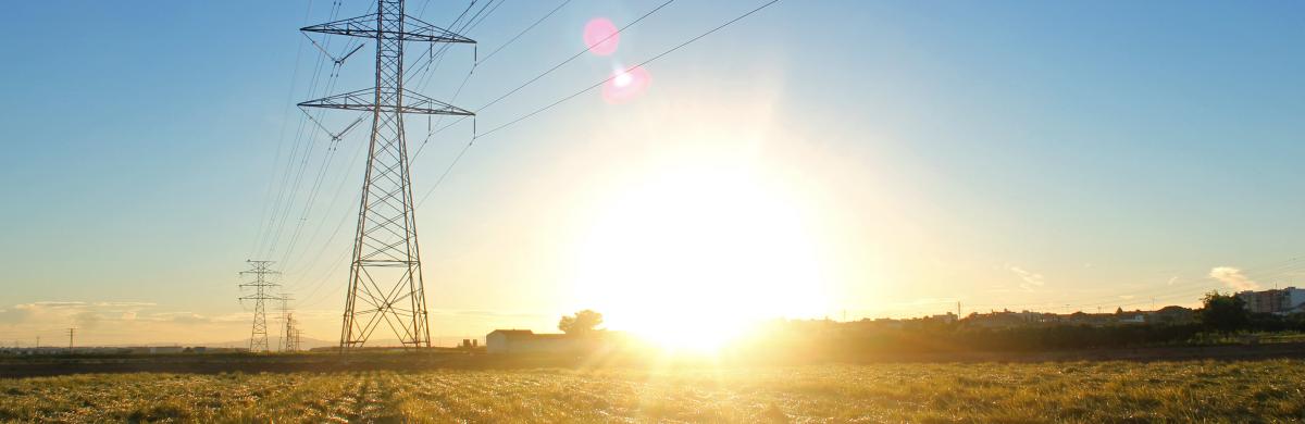 A field landscape with electricity pylons and overhead cabling and a bright sun setting in a blue sky 